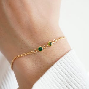 May include: A gold bracelet with three gemstones: two green and one olive-green. The bracelet has a fine chain design, with the gemstones set in a row. The bracelet is shown on a wrist, with a white sweater in the background.