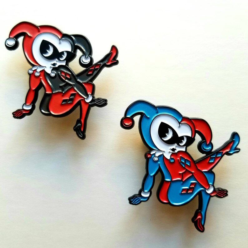 Puede incluir: Dos pines de esmalte con un personaje de dibujos animados con ropa roja y negra y un sombrero de buf&oacute;n. Un pin es rojo y negro, el otro es azul y rojo.