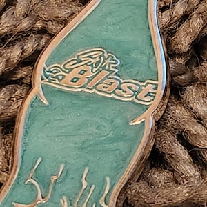 May include: A teal-colored enamel pin in the shape of a bottle. The pin features the word "Blast" in a gold, stylized font. The pin has a gold-colored border and is set against a textured, brown background.