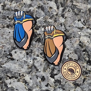May include: Two enamel pins featuring a cartoon fist with a mechanical hand. The fist on the left is blue and yellow, while the fist on the right is brown and yellow. The pins are on a gray and brown speckled surface. The text "SPECIAL VARIANT" is on a brown circle with a star on each side.