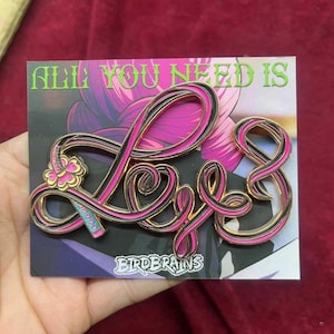 May include: A gold and pink enamel pin with the word "Love" in a stylized font. The pin has a black outline and a small floral design on the left side. The text "All you need is" is printed above the word "Love" and the text "Birdbrains" is printed below the word "Love".