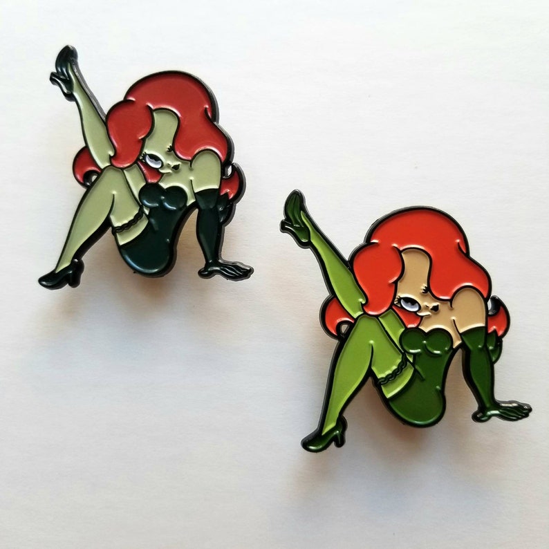 Puede incluir: Dos pines de esmalte que representan a un personaje de dibujos animados con cabello rojo, ropa verde y accesorios negros. El personaje est&aacute; en una pose sugerente.