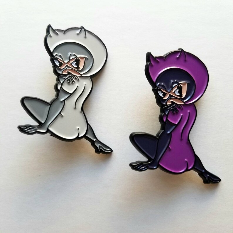 Puede incluir: Dos pines de esmalte con un personaje de dibujos animados con orejas de gato y una m&aacute;scara. Un pin es blanco con detalles negros, y el otro es morado con detalles negros.