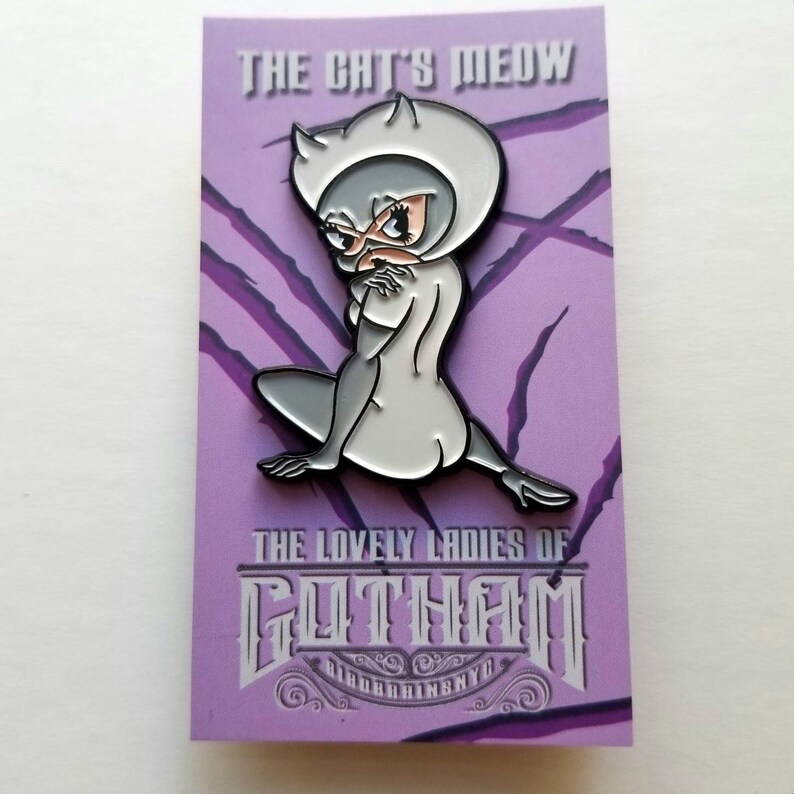 Puede incluir: Un pin de esmalte plateado con una representaci&oacute;n de dibujos animados de Catwoman, un personaje ficticio de los c&oacute;mics de Batman. El pin est&aacute; sobre un fondo morado con el texto "The Cat's Meow" y "The Lovely Ladies of Gotham" en una fuente de estilo vintage.