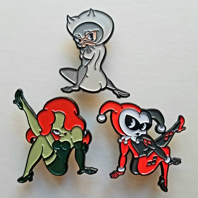 Puede incluir: Tres pines de esmalte con personajes de dibujos animados. El primer pin es un gato gris con la cara blanca y los ojos negros. El segundo pin es una mujer de piel verde con el pelo rojo y un atuendo negro. El tercer pin es un personaje rojo y negro con un gorro de buf&oacute;n y la cara blanca.