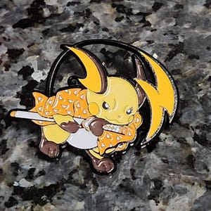 May include: A yellow and orange enamel pin featuring a cartoon Pikachu dressed as a demon slayer. The pin has a black outline and a silver lightning bolt design.