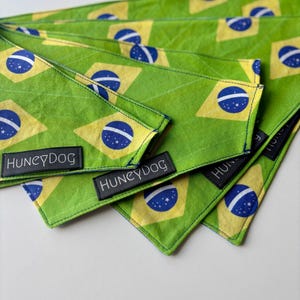 Puede incluir: Bandanas verdes para perros con el diseño de la bandera brasileña, con un rombo amarillo con un círculo azul y estrellas blancas. Cada bandana tiene una etiqueta negra que dice "HUNEYDOG". Las bandanas están hechas de tela texturizada.