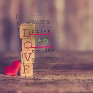May include: A clear acrylic wedding invitation with the words "YOU ARE INVITED TO THE MARRIAGE OF" at the top. The names "Jaclyn + Brandon" are spelled out with wooden blocks stacked on top of each other. The words "LOVE" are spelled out with wooden blocks below the names. The invitation also includes the date, time, and location of the wedding.