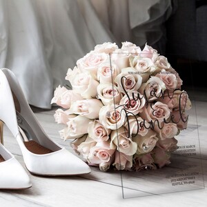 May include: A clear acrylic wedding invitation with the names "Susan & Daniel" written in black calligraphy. The invitation is placed on a wooden surface with a bouquet of blush pink roses and a pair of white high heels.