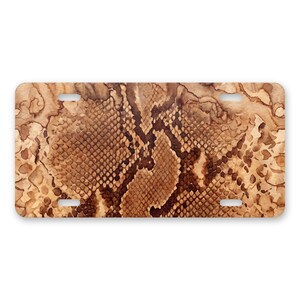 May include: A rectangular license plate featuring a brown and tan snakeskin pattern. The design displays a detailed, repeating scale texture across the entire surface, with four mounting holes positioned near the corners of the plate.