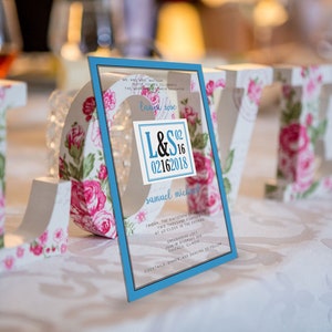 May include: A blue and white wedding invitation with the text "L&S 02 16 2018" and "Samuel Michael" on a white background. The invitation is sitting on a table with a floral design.