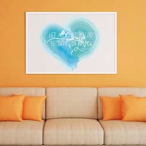 May include: A framed art print with a white border, featuring a blue watercolor heart with the words "LET ALL THAT YOU DO BE DONE IN Love." The print hangs above a beige sofa with orange throw pillows.