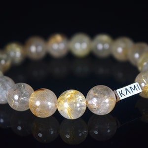 Op de afbeelding: Een armband van gouden en helder kwarts met een zilveren label waarop "KAMI" staat.