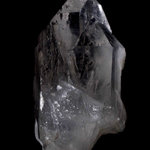 Puede incluir: Un cristal de cuarzo transparente sobre un fondo negro. El cristal tiene una punta afilada y una base rugosa y texturizada. Las facetas del cristal reflejan la luz, creando un efecto brillante. Un espécimen geológico natural.