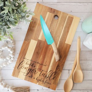 May include: A wooden chopping board with a teal knife and wooden spoons. The board has a natural wood grain pattern and the text "when you know more than you and build a longer table not a higher fence."