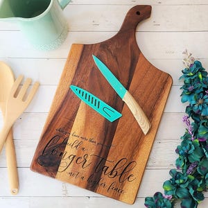 May include: A wooden chopping board with a handle, a teal knife, and a teal knife sheath. The cutting board has the text "when you have more than you need, build a longer table not a higher fence." Wooden spoon and fork are on the left.