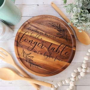 May include: A round wooden serving board with the engraved text "when you have more than you need, build a longer table not a higher fence." The board is surrounded by wooden utensils and decorative elements.