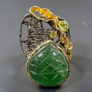Könnte beinhalten: Ein Ring mit einem grünen Edelstein mit einem geschnitzten Design, der in einer filigranen Fassung aus Gold und Silber gefasst ist. Der Ring hat zwei kleinere grüne Edelsteine.