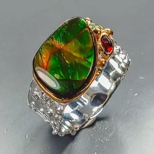Op de afbeelding: Een ring met een grote, iriserende groene en oranje ammolietsteen in een gouden zetting. De band is gemaakt van getextureerd, gehamerd zilver en bevat kleine decoratieve gouden accenten samen met een enkele, kleine, ronde, donkerrode edelsteen.