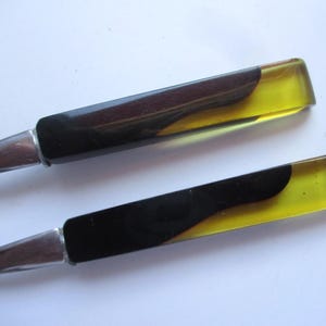 Può includere: Due aprilettere vintage con lame e manici in metallo argentato. I manici presentano un design nero, marrone e giallo. Gli aprilettere sono su uno sfondo bianco.
