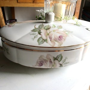 Könnte beinhalten: Weißes Porzellan-Serviergeschirr mit Deckel, verziert mit einem floralen Rosenmuster in Rosa und Grün. Das Geschirr hat goldfarbene Verzierungen und Griffe sowie einen passenden Deckel mit einem kleinen Knauf. Ein klassisches Stück zum Servieren von Speisen.