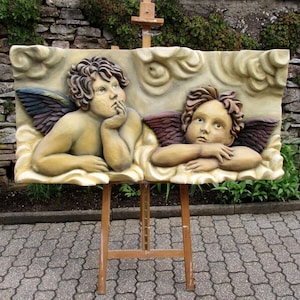 May include: A wooden sculpture of two cherubs with wings, painted in shades of brown, gold, and white, against a background of clouds. The sculpture is displayed on an easel.