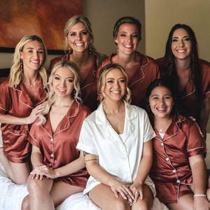 May include: A group of seven people wearing matching rust-colored pajamas with white piping. The person in the center is wearing a white pajama top. They are all smiling and looking at the camera.