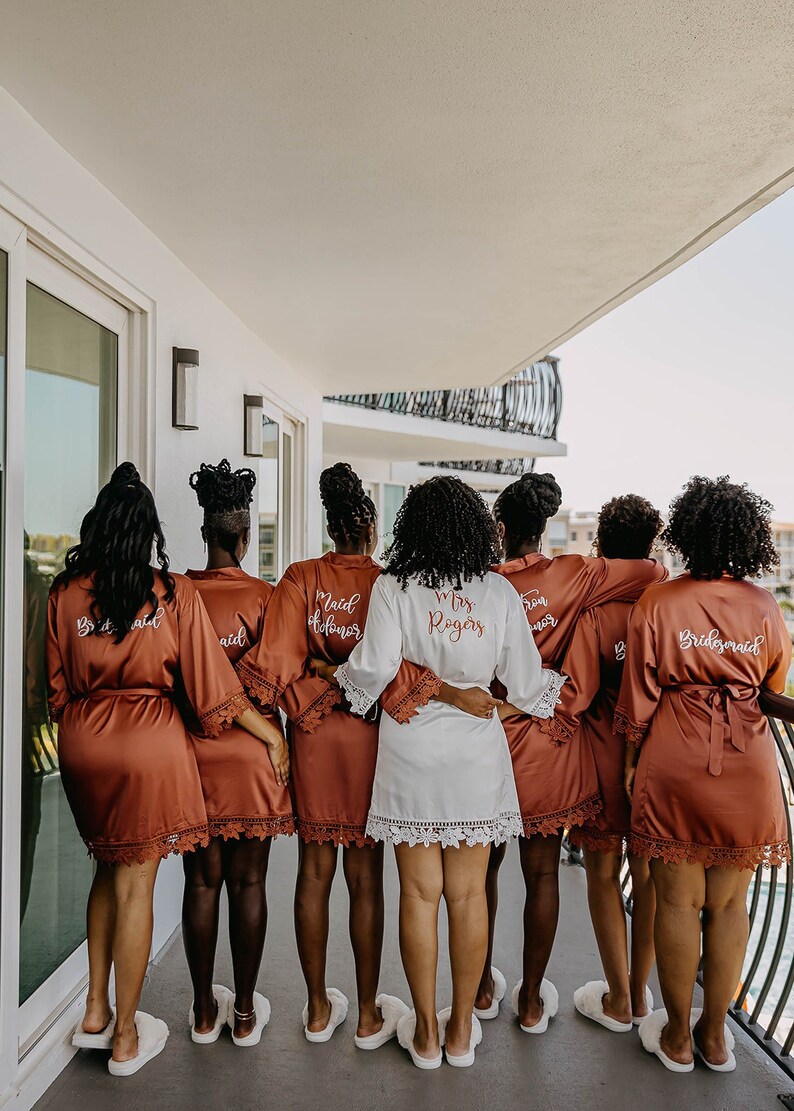 Op de afbeelding: Een groep van acht mensen die bijpassende gewaden dragen met geborduurde tekst. De gewaden zijn roestkleurig met witte kantversiering. De tekst op de gewaden luidt "Bride", "Maid of Honor", "Mrs. Rogers", "Bridesmaid".