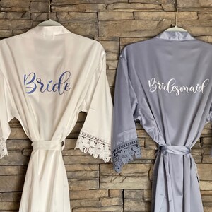 Op de afbeelding: Twee satijnen badjassen met kantversiering, een witte met "Bride" geborduurd in blauw, de andere grijs met "Bridesmaid" geborduurd in wit.