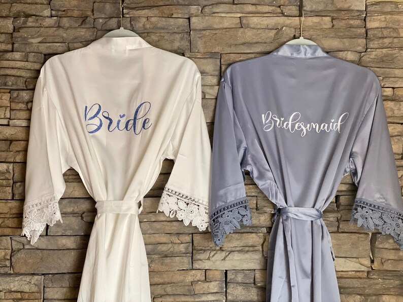 Op de afbeelding: Twee satijnen badjassen met kantversiering, een witte met "Bride" geborduurd in blauw, de andere grijs met "Bridesmaid" geborduurd in wit.