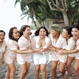 May include: A group of women, including a bride, laugh together on a beach. The women wear matching light pink pajama sets, while the bride wears a white dress. The Creative Skills logo is in the bottom left corner.
