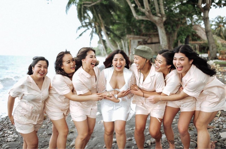 May include: A group of women, including a bride, laugh together on a beach. The women wear matching light pink pajama sets, while the bride wears a white dress. The Creative Skills logo is in the bottom left corner.