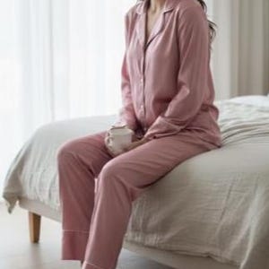 May include: A light pink pajama set with white trim, consisting of a button-down shirt and pants. The person is wearing white open-toe slippers and holding a white mug. The pajamas are made of a soft, silky material.