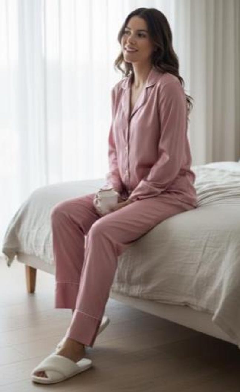 May include: A light pink pajama set with white trim, consisting of a button-down shirt and pants. The person is wearing white open-toe slippers and holding a white mug. The pajamas are made of a soft, silky material.