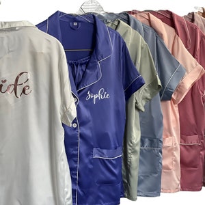 May include: A row of satin pajama sets in various colors, including white, blue, pink, and beige, hang on a rack. The white set has "Bride" printed on the back in rose gold. The blue set has "Sophie" embroidered on the front.