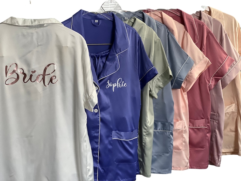 May include: A row of satin pajama sets in various colors, including white, blue, pink, and beige, hang on a rack. The white set has "Bride" printed on the back in rose gold. The blue set has "Sophie" embroidered on the front.
