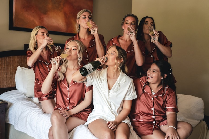 May include: A group of people in matching rust-colored satin pajamas and white pajamas, toasting with champagne flutes and a champagne bottle. The scene is set in a bedroom with a bed and a painting on the wall.