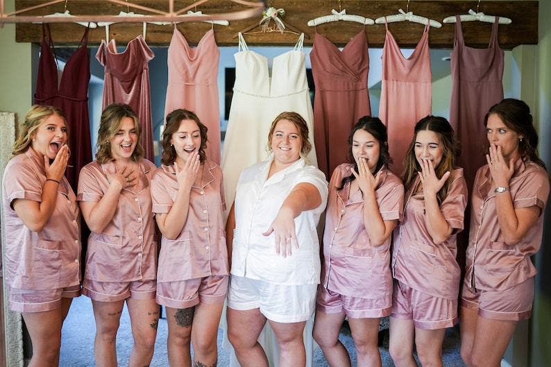 May include: A group of women in matching pink pajama sets and a bride in white pajamas, all reacting with surprise. A wedding dress and bridesmaid dresses hang in the background, suggesting a bridal party. The image captures a moment of excitement.