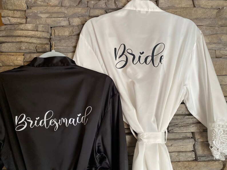 Op de afbeelding: Twee satijnen badjassen, een zwarte en een witte, met de woorden "Bride" en "Bridesmaid" geborduurd op de rug.