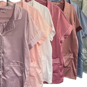 May include: A row of silk pajama sets in various colors, including mauve, pink, white, and blue. Each set features a button-down top with a notched collar, short sleeves, and a chest pocket, all trimmed with white piping.