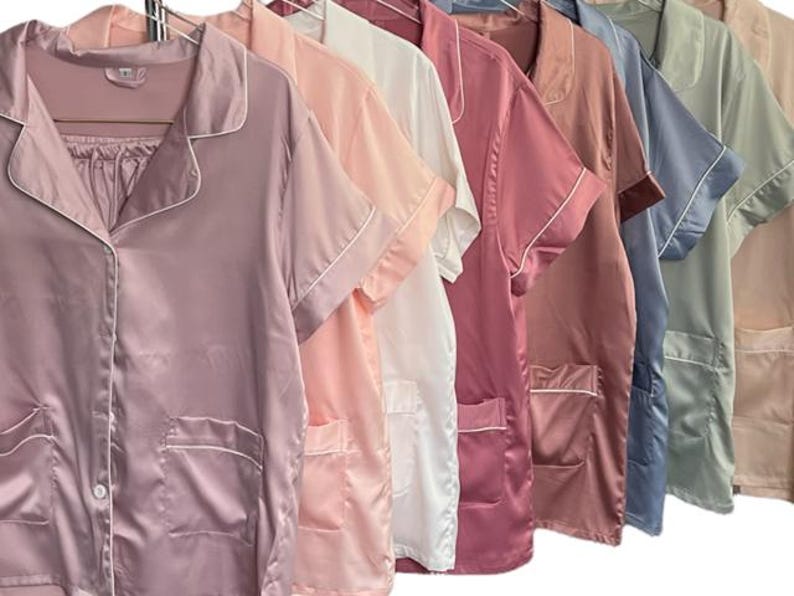 May include: A row of silk pajama sets in various colors, including mauve, pink, white, and blue. Each set features a button-down top with a notched collar, short sleeves, and a chest pocket, all trimmed with white piping.