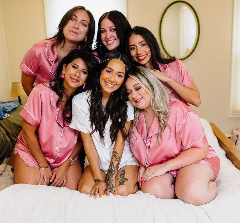 May include: A group of six people, smiling and posing on a bed. Five are wearing matching pink satin pajama sets with white trim. One person is wearing a white button-down shirt. The room has a neutral color palette.