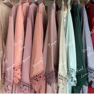Op de afbeelding: Een rij van 12 badjassen in verschillende kleuren, waaronder zachte lila, wit, champagne, stoffig roze, mauve, rozenhout, taupe, Tiffany blauw, bosgroen, stoffig blauw, wijn, marine, kaneel en stoffig koraal. Elke badjas heeft een kantversiering rond de zoom en de mouwen.