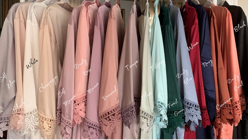 Op de afbeelding: Een rij van 12 badjassen in verschillende kleuren, waaronder zachte lila, wit, champagne, stoffig roze, mauve, rozenhout, taupe, Tiffany blauw, bosgroen, stoffig blauw, wijn, marine, kaneel en stoffig koraal. Elke badjas heeft een kantversiering rond de zoom en de mouwen.
