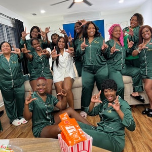 May include: A group of people wearing matching green satin pajamas with white piping, some holding up their hands in a letter formation. A popcorn container with orange packets is on the table. The room has a ceiling fan and a neutral color scheme.