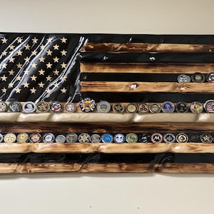 May include: Wooden American flag with a distressed finish, featuring a black stripe and rows for displaying challenge coins. The flag has stars in the upper left corner and multiple challenge coins displayed on the shelves.