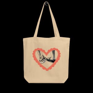 Op de afbeelding: Beige draagtas met een rood hartvormig ontwerp. Het hart bevat een zwart-wit illustratie van twee paar voeten in schoenen. Eén voet draagt een zwart-witte schoen, de andere een zwarte schoen.