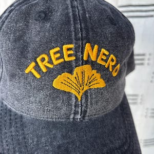 Könnte beinhalten: Dunkelgraue Baseballkappe mit dem gestickten Text "TREE NERD" in Gelb sowie einem gelben Ginkgoblatt-Design. Die Kappe besteht aus einem strukturierten Stoff und hat einen gebogenen Schirm.