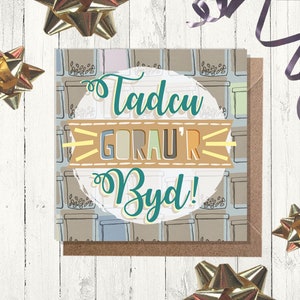 May include: A greeting card with a white background and a circle in the center. The circle has a light blue border and contains the text "Tadcu Gorau'r Byd!" in a colorful font.