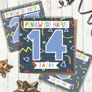 Puede incluir: Una tarjeta de cumpleaños con un diseño colorido de estilo años 90. La tarjeta presenta el número 14 en azul con el nombre Jacob debajo. El texto "Penblwydd Hapus" está escrito en galés en la parte superior de la tarjeta.